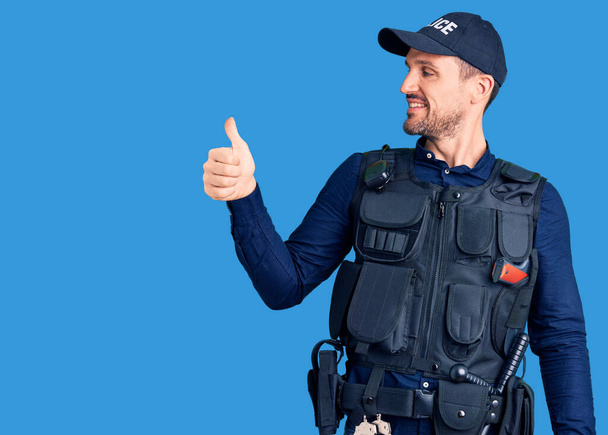 Young handsome man wearing police uniform looking proud, smiling doing thumbs up gesture to the side  - Photo, Image