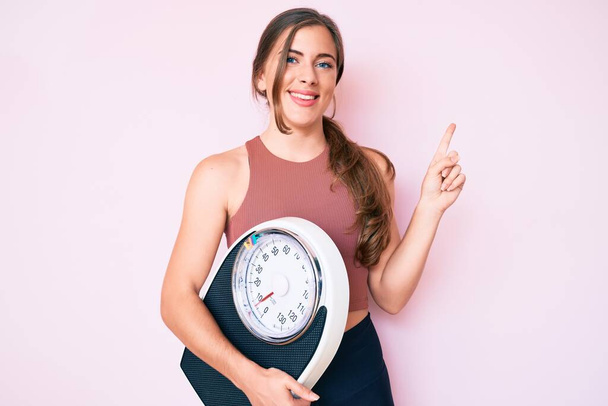 Schöne junge Kaukasierin hält Gewichtsmaschine, um Gewichtsverlust auszugleichen lächelnd glücklich zeigt mit Hand und Finger zur Seite  - Foto, Bild