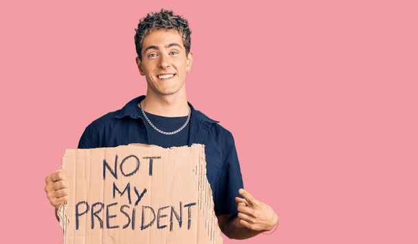 Junger gutaussehender Mann hält nicht mein Präsident Protestbanner und lächelt glücklich und zeigt mit Händen und Fingern  - Foto, Bild