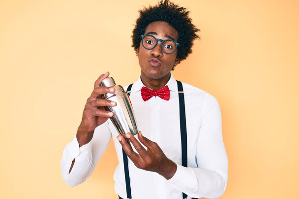Handsome african american bartender man with afro hair preparing cocktail mixing drink with shaker looking at the camera blowing a kiss being lovely and sexy. love expression.  - 写真・画像
