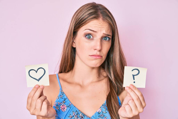 Young blonde girl holding heart and question mark reminder clueless and confused expression. doubt concept.  - Photo, Image