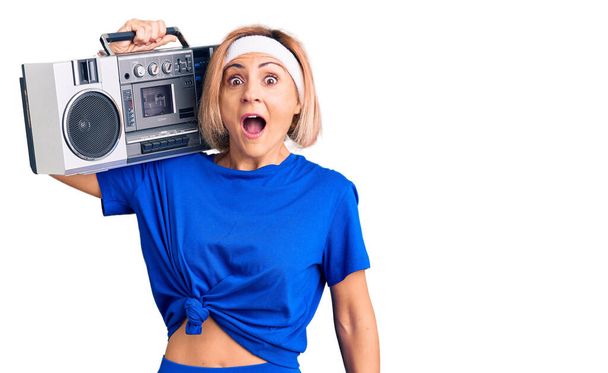 Young blonde woman wearing sportswear holding boombox, listening to music scared and amazed with open mouth for surprise, disbelief face  - Fotó, kép