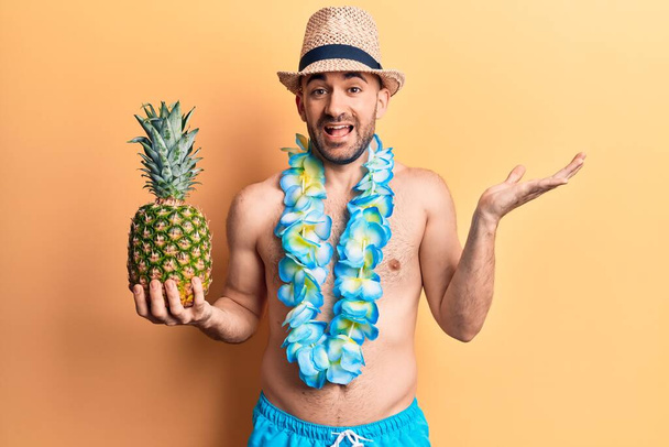 Молодой красивый лысый мужчина в купальниках и гавайских леях с ананасом в руках празднует достижение со счастливой улыбкой и выражением победителя с поднятой рукой  - Фото, изображение