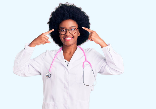 Mladá Afroameričanka v lékařském kabátě a stetoskopu s úsměvem ukazujícím na hlavu s oběma prsty, skvělý nápad nebo myšlenka, dobrá paměť  - Fotografie, Obrázek