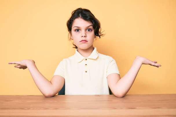 Aranyos spanyol gyerek alkalmi ruhát visel ül az asztalon tanácstalan és zavaros kifejezés a karok és a kezek fel. kétség.  - Fotó, kép