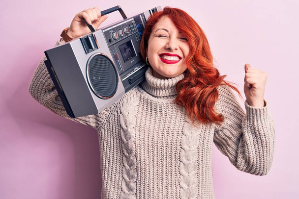 Gyönyörű vörös hajú nő hallgat zenét gazdaság vintage boombox felett rózsaszín háttér sikoltozó büszke, ünneplő győzelem és a siker nagyon izgatott felemelt karral - Fotó, kép