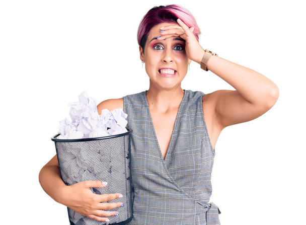 Молодая красивая женщина с розовыми волосами, держащая в руках бумажную корзину, полную скомканных бумаг, напряженная и разочарованная рукой на голове, удивленная и сердитая  - Фото, изображение