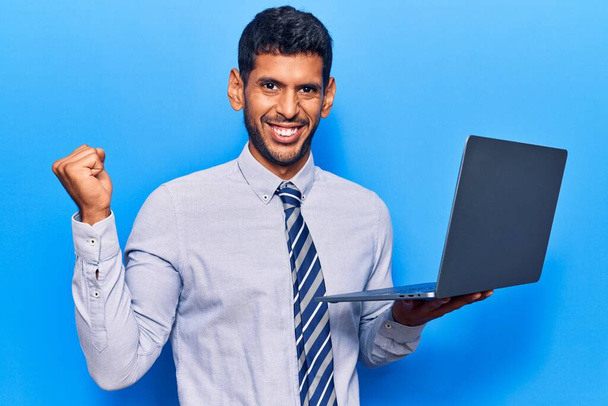 Joven latino sosteniendo laptop celebrando victoria con sonrisa feliz y expresión ganadora con las manos levantadas  - Foto, Imagen