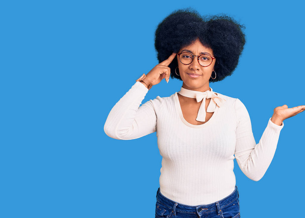 カジュアルな服と眼鏡を身に着けている若いアフリカ系アメリカ人の少女は、コピースペースと額に指を指すことを示すオープン手のひらに混乱し、悩まされている。考えてみてください.  - 写真・画像