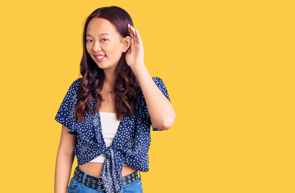 Menina chinesa bonita nova vestindo roupas casuais sorrindo com a mão sobre o ouvido ouvindo um rumor ou fofoca. conceito de surdez.  - Foto, Imagem