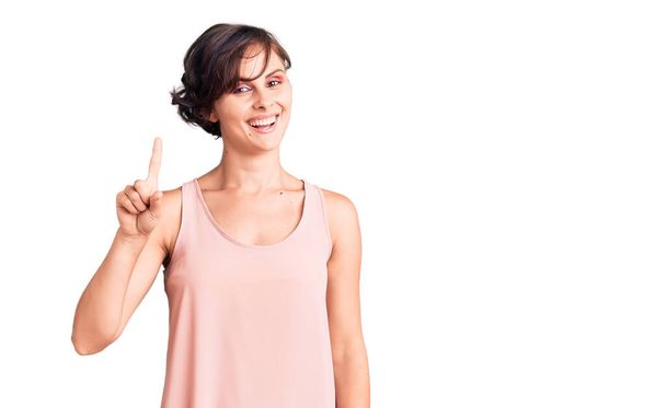 Beautiful young woman with short hair wearing casual style with sleeveless shirt showing and pointing up with finger number one while smiling confident and happy.  - Fotografie, Obrázek