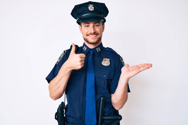 Молодой кавказский мужчина в полицейской форме показывает ладонь и делает нормальный жест с большими пальцами вверх, улыбаясь счастливой и веселой  - Фото, изображение