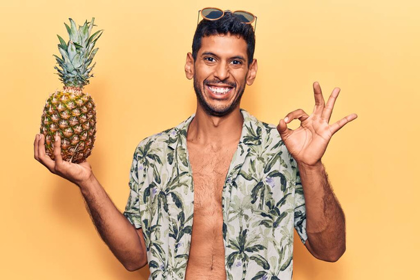 Молодий латинський чоловік у літній сорочці тримає ананас, робить знак з пальцями, усміхнений дружній жест відмінний символ
  - Фото, зображення