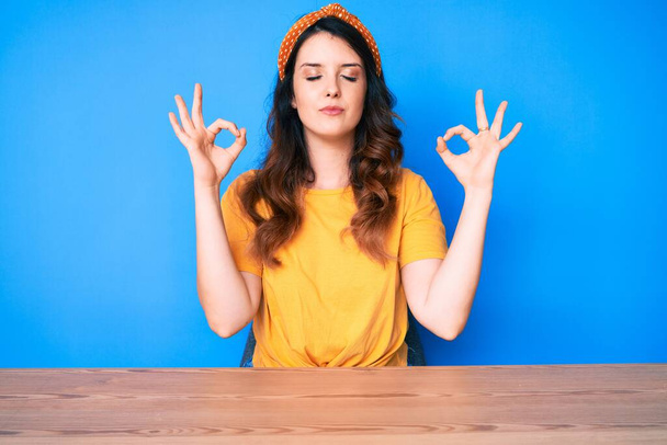 Young beautiful brunette woman sitting on the table over blue background relax and smiling with eyes closed doing meditation gesture with fingers. yoga concept.  - Photo, Image