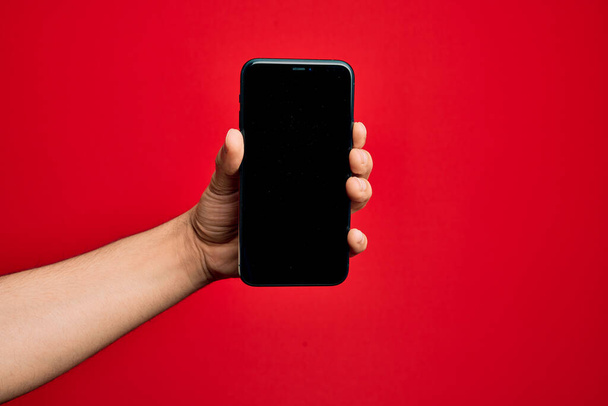 Hand of caucasian young man holding smartphone showing screen over isolated red background - Fotoğraf, Görsel