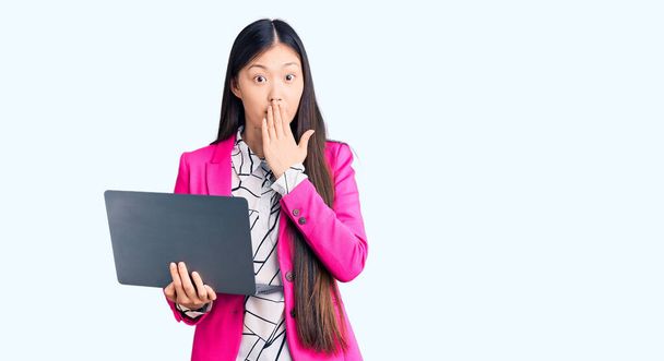 Молодая красивая китаянка, использующая ноутбук, прикрывающий рот рукой, шокирована и боится ошибиться. Удивительное выражение  - Фото, изображение