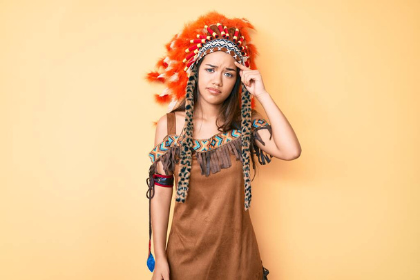Mladá krásná latinská dívka v indiánském kostýmu ukazující nešťastně na pupínek na čele, ošklivá infekce hlavy. akné a kožní problémy  - Fotografie, Obrázek
