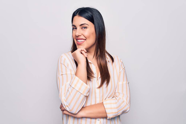 Young beautiful brunette woman wearing casual striped shirt standing over white background smiling looking confident at the camera with crossed arms and hand on chin. Thinking positive. - Foto, afbeelding