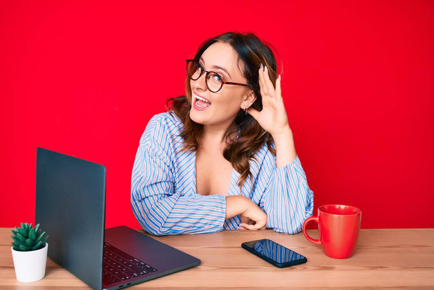Jonge mooie blanke vrouw die op kantoor werkt met een laptop lachend met hand op hand oor luisterend en horend naar geruchten of roddels. doofheid.  - Foto, afbeelding