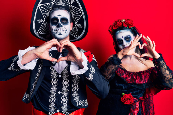 Pareja joven vistiendo el día mexicano del disfraz muerto sobre rojo sonriendo en el amor haciendo forma de símbolo del corazón con las manos. concepto romántico.  - Foto, imagen