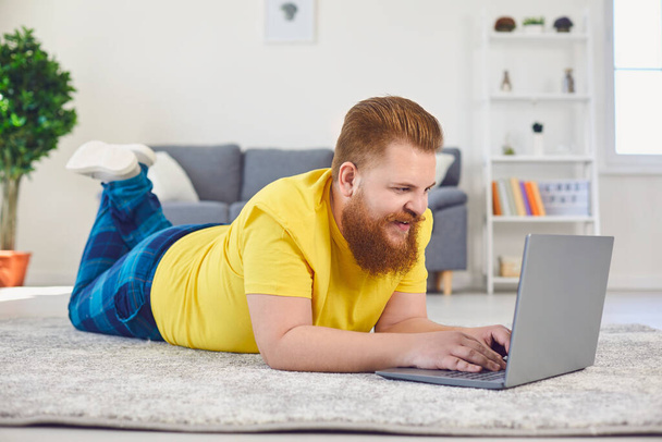 Online job training communication date. Funny man with a beard is typing in a laptop while lying on the floor at home - Foto, Bild
