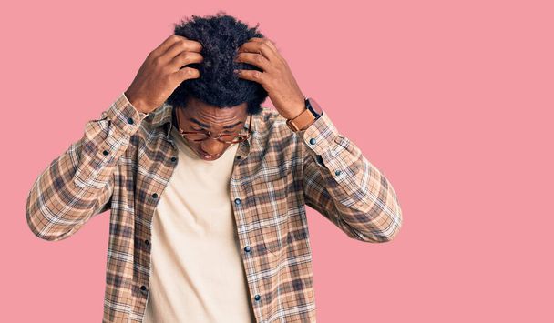 アフロヘアのハンサムなアフリカ系アメリカ人男性は、痛みや片頭痛のために絶望的な頭痛やストレスに苦しんでカジュアルな服や眼鏡を着用しています。手を頭に.  - 写真・画像