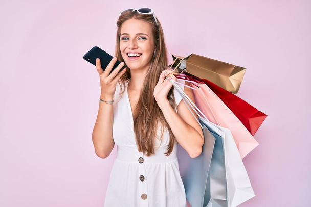 Молодая блондинка держит сумки с покупками разговаривая по смартфону улыбаясь с счастливой и прохладной улыбкой на лице. показывая зубы.  - Фото, изображение