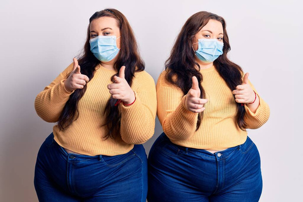 Young plus size twins wearing medical mask pointing fingers to camera with happy and funny face. good energy and vibes.  - Photo, Image