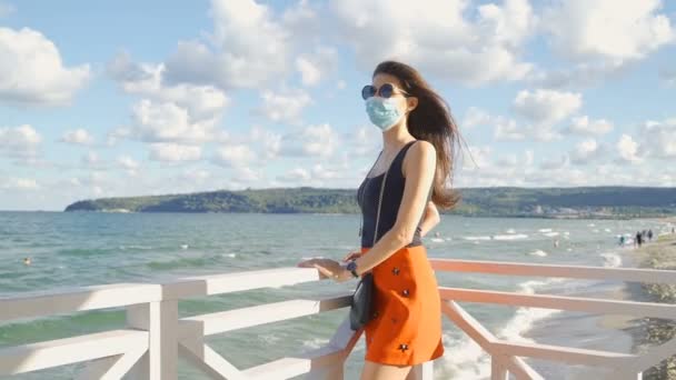 Μια γυμνασμένη γυναίκα με γυαλιά ηλίου και μάσκα προσώπου που κοιτάει πάνω από ένα φράχτη στη θάλασσα. Μέτρα προστασίας κατά την πανδημία - Πλάνα, βίντεο