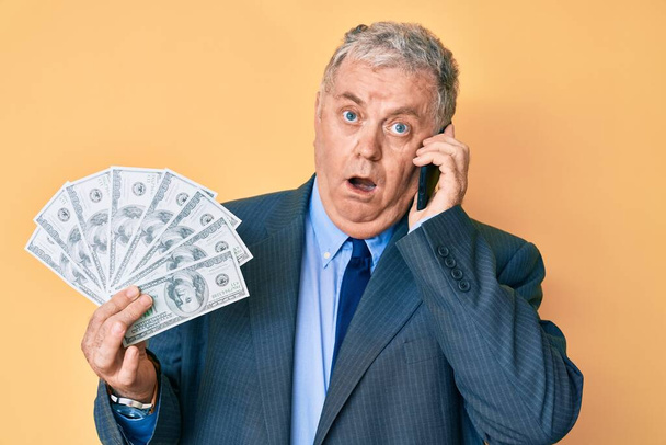 Älterer grauhaariger Mann im Anzug, Dollar in der Hand und Smartphone im Schockgesicht, sieht skeptisch und sarkastisch aus, überrascht mit offenem Mund  - Foto, Bild