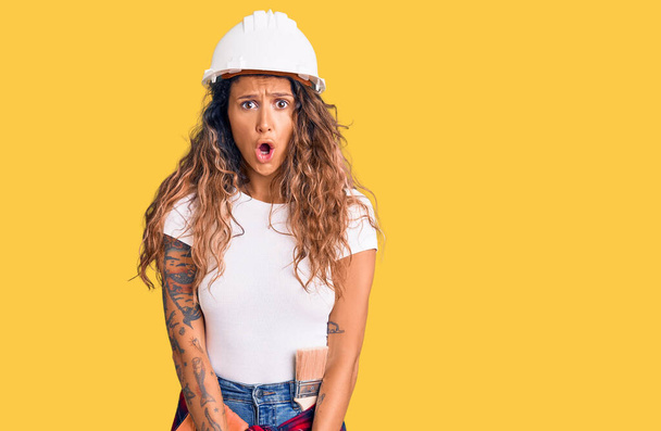 Young hispanic woman with tattoo wearing hardhat and builder clothes afraid and shocked with surprise expression, fear and excited face.  - Photo, Image