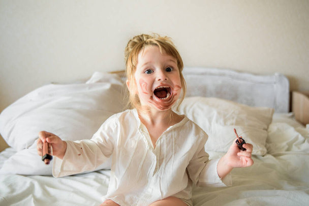 Little girl doing makeup daughter have fun. The baby paints lips. Mom's cosmetics.The child is having fun on the bed at home - Foto, imagen
