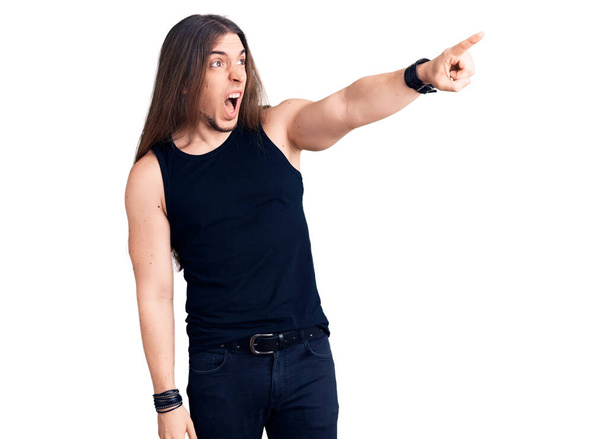 Young adult man with long hair wearing goth style with black clothes pointing with finger surprised ahead, open mouth amazed expression, something on the front  - Foto, imagen