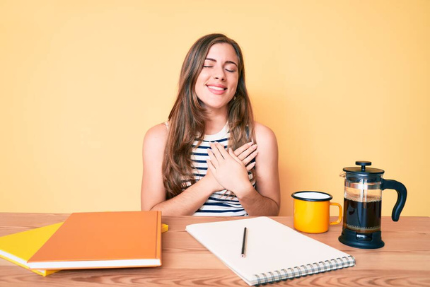 Beautiful young caucasian woman sitting on the table stuying for university smiling with hands on chest, eyes closed with grateful gesture on face. health concept.  - Фото, изображение