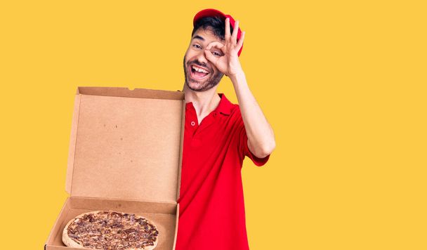 Junger hispanischer Mann hält Lieferung Pizzaschachtel lächelnd glücklich tut ok Zeichen mit Hand auf Auge durch Finger schauen  - Foto, Bild