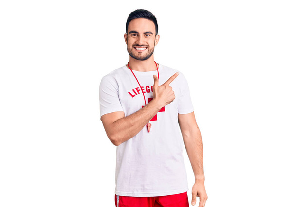 Young handsome man wearing lifeguard uniform cheerful with a smile of face pointing with hand and finger up to the side with happy and natural expression on face  - Photo, Image