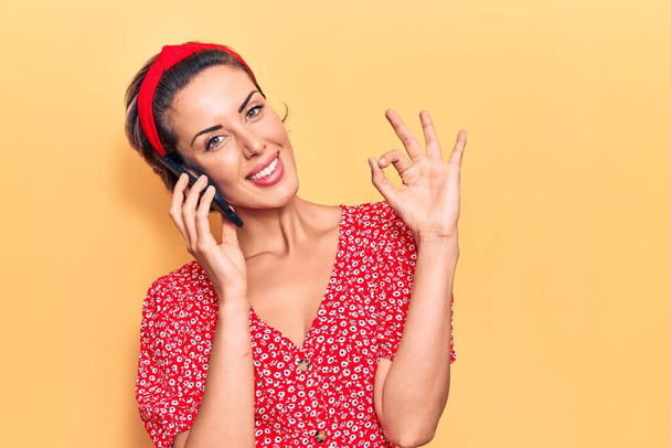 Young beautiful woman having conversation talking on the smartphone doing ok sign with fingers, smiling friendly gesturing excellent symbol  - Fotografie, Obrázek