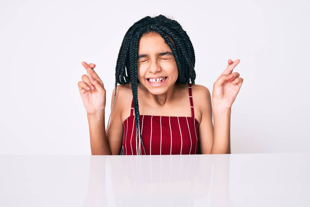 Young african american girl child with braids wearing casual clothes sitting on the table gesturing finger crossed smiling with hope and eyes closed. luck and superstitious concept.  - Foto, immagini