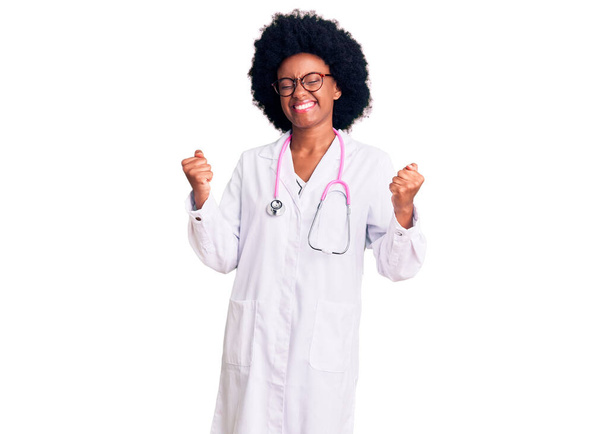 Молода афроамериканська жінка, одягнена в лікарське пальто і стетоскоп, дуже щаслива і захоплена жестом переможця з піднятими руками, посміхається і кричить про успіх. Святкування.  - Фото, зображення