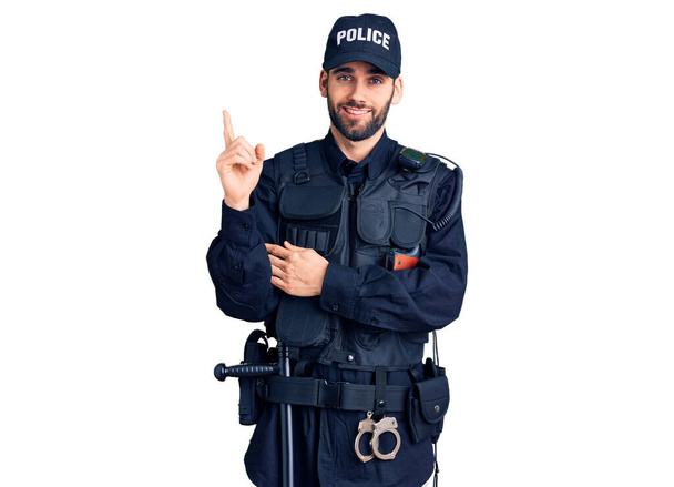 Sakallı, polis üniforması giyen, yüzünde kocaman bir gülümseme olan, eli ve parmağı kameraya bakan genç yakışıklı bir adam..  - Fotoğraf, Görsel