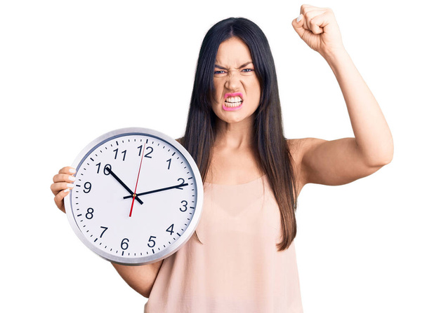 Young beautiful caucasian woman holding big clock annoyed and frustrated shouting with anger, yelling crazy with anger and hand raised  - Photo, image