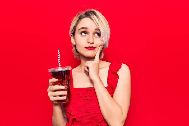 Fiatal gyönyörű szőke nő iszik pohár kóla ital komoly arc gondolkodás kérdés kézzel az állon, gondolkodó zavaros ötlet  - Fotó, kép