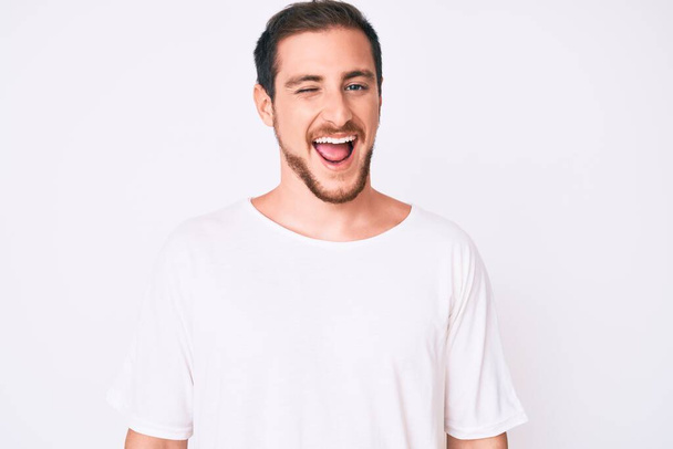 Young handsome man wearing casual white tshirt winking looking at the camera with sexy expression, cheerful and happy face.  - Φωτογραφία, εικόνα
