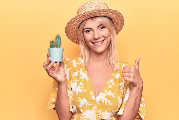 美しいブロンド女性身に着けている夏の帽子保持サボテン植物ポット以上黄色の背景笑顔幸せと正、親指アップ行う優れた承認サイン - 写真・画像