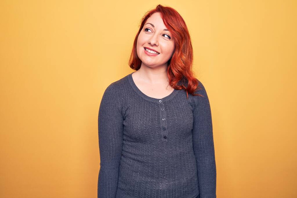 Young beautiful redhead woman wearing casual sweater standing over yellow background looking to side, relax profile pose with natural face and confident smile. - Photo, Image