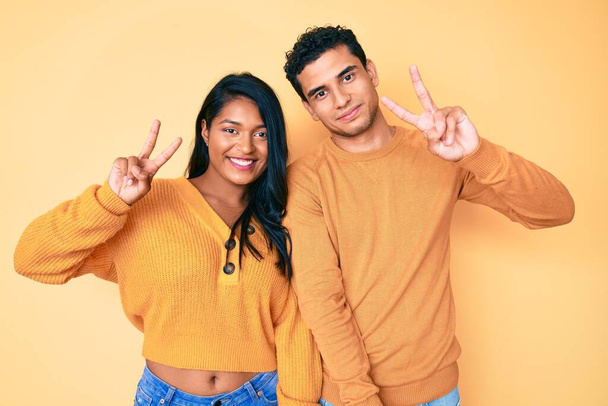 Belo casal jovem latino vestindo roupas casuais juntos sorrindo olhando para a câmera mostrando os dedos fazendo sinal de vitória. Número dois..  - Foto, Imagem