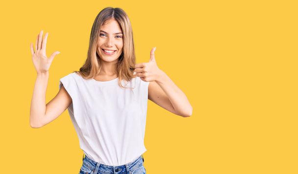 Beautiful caucasian woman with blonde hair wearing casual white tshirt showing and pointing up with fingers number six while smiling confident and happy.  - Foto, immagini