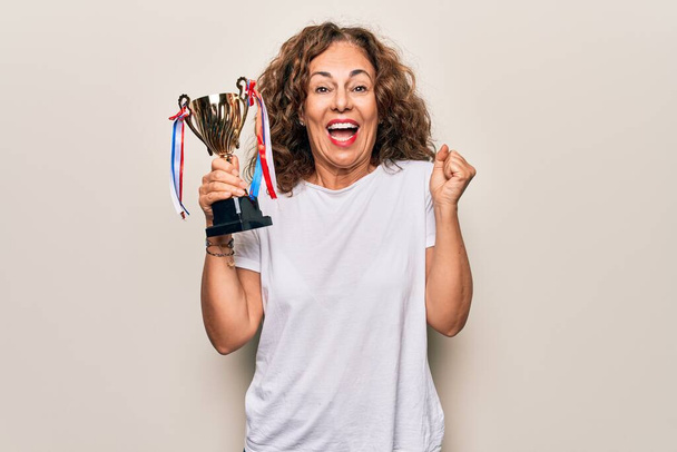 Orta yaşlı başarılı bir kadın zafer kupasını beyaz arka planda tutarken gururla haykırıyor, zaferi ve başarıyı kutluyor. - Fotoğraf, Görsel