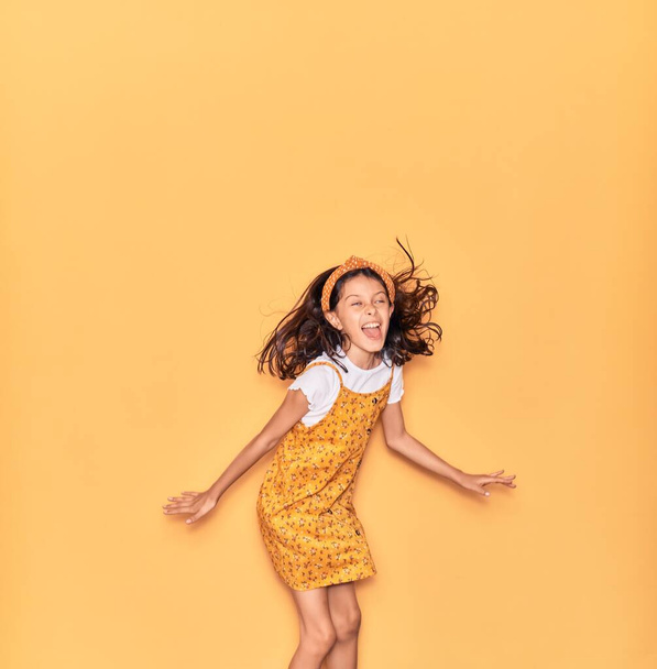 Adorable hispanic child girl wearing casual dress and diadem smiling happy. Jumping with smile on face over isolated yellow background - Photo, image