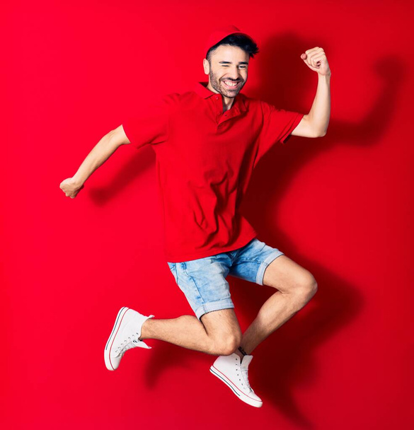 Young handsome deliveryman wearing cap smiling happy. Jumping with smile on face over isolated red background - Foto, afbeelding